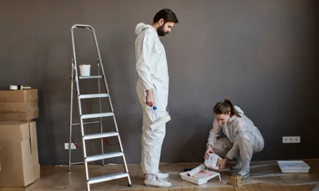 Pareja lista para pintar con pintura. Él sostiene un rodillo, ella vierte pintura en una bandeja. Hay cajas y una escalera.
