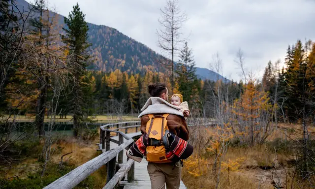 Ojciec z dzieckiem na plecach spaceruje drewnianą kładką pośród jesiennych lasów. Tatry z dzieckiem to piękna przygoda.