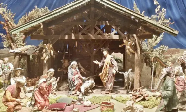Krippenszene mit Maria, Josef und dem Jesuskind. Engel schweben über der Szene, die an Weihnachten im Mittelalter erinnert. Hirten und Musiker sind versammelt.