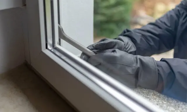 Manos enguantadas forzando una ventana de seguridad con una palanca.