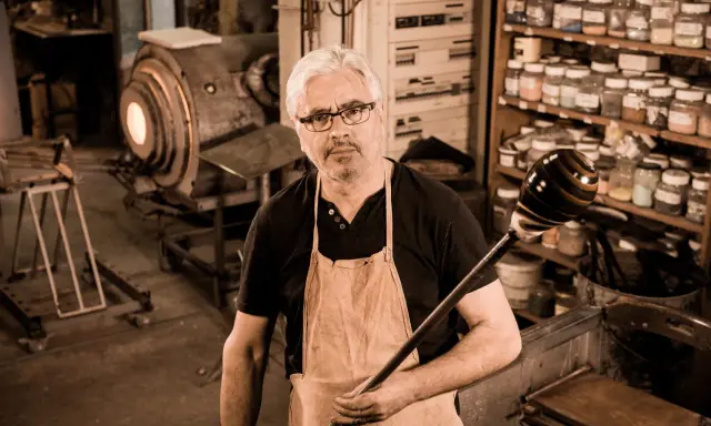Un artisan verrier expérimenté, tablier en cuir, tient sa canne à souffler devant son four incandescent.