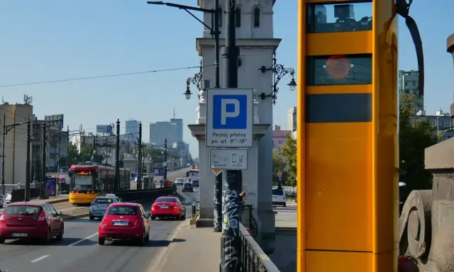 Żółty fotoradar stoi przy drodze, gdzie widać ruch samochodowy i tramwaj. Obok znak parkingu płatnego.