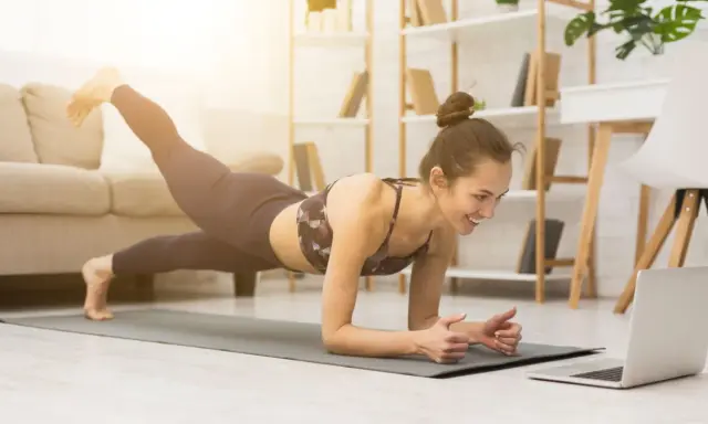 Uśmiechnięta kobieta w desce z nogą w górze, oglądająca trening na laptopie. To świetny sposób, jak zmotywować się do ćwiczeń w domu.