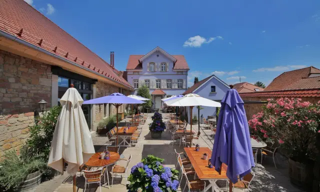 Terrasse eines Weinguts mit Tischen, Stühlen, Sonnenschirmen und Blumen unter blauem Himmel.