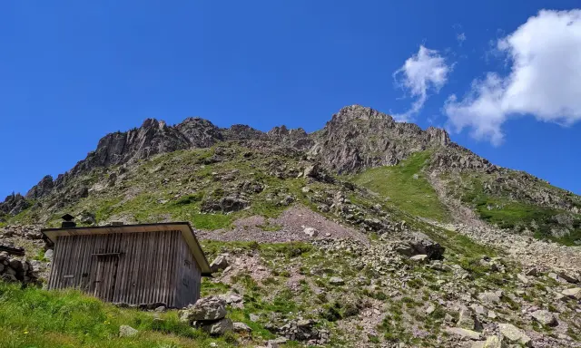 Bivacco Paolo e Nicola, un rifugio in legno ai piedi di montagne rocciose sotto un cielo azzurro.