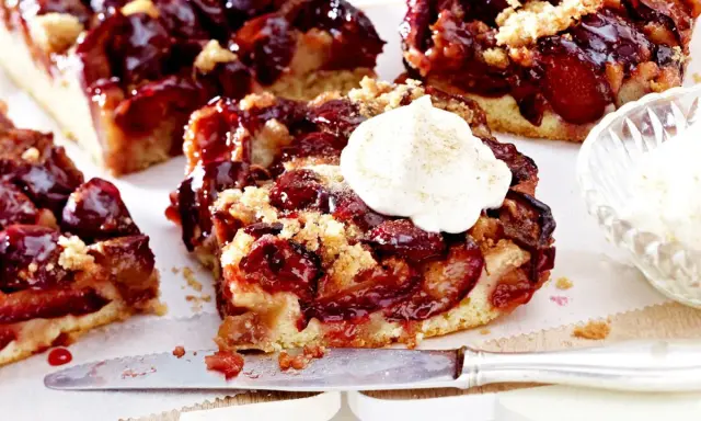 Saftiger Pflaumenkuchen mit Streuseln und einem Klecks Sahne. Ein Stück Kuchen liegt auf einem Messer, daneben eine Schale mit Sahne.