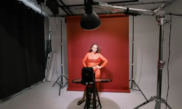 Modelka w pomarańczowej sukience pozuje na tle czerwonego tła. Wokół niej profesjonalne **światło studyjne** i sprzęt fotograficzny.