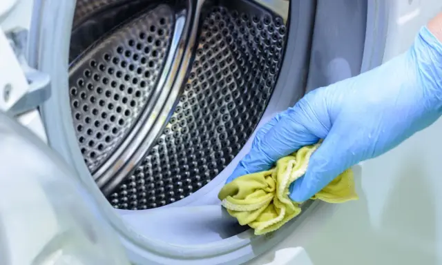 Mano con guante azul limpiando el borde de la lavadora con un paño amarillo. Un método natural para limpiar lavadora vinagre y bicarbonato.