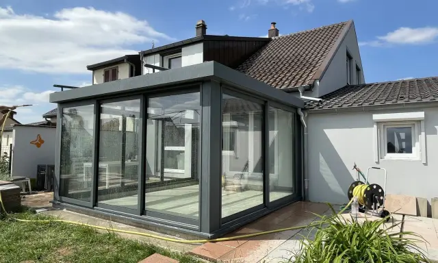 Une véranda moderne en verre et métal, prête pour l'assurance. Le soleil brille sur le jardin et la maison.