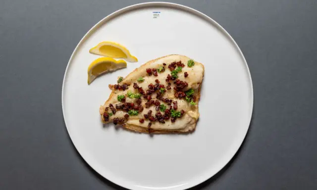 Scholle Finkenwerder Art: gebratenes Fischfilet mit Speckwürfeln und Petersilie, garniert mit Zitronenspalten auf einem weißen Teller.
