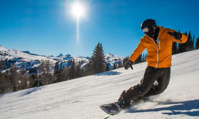 Snowboardzista w pomarańczowej kurtce zjeżdża po ośnieżonym stoku w słoneczny dzień.