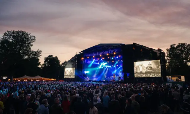 Stor publik vid Rosendal Garden Party. En scen med blå ljus och artister, omgiven av träd och en vacker kvällshimmel.