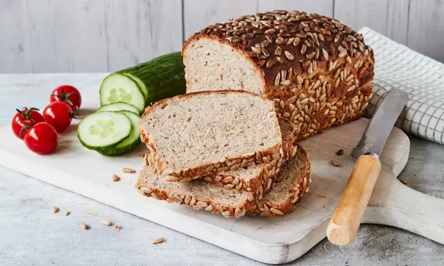 Frisch gebackenes Sonnenblumenbrot, zubereitet im Thermomix, liegt auf einem Holzbrett neben Tomaten und Gurkenscheiben.