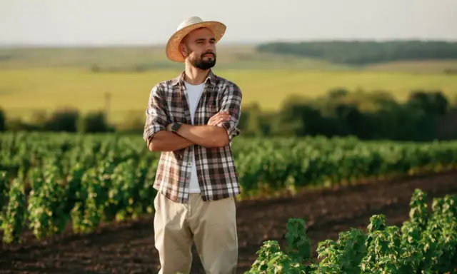 Rolnik w słomkowym kapeluszu, z rękami skrzyżowanymi na piersi, patrzy w dal na swoje pole. Myśli o przyszłości i o tym, jak krus a umowa zlecenie wpłyną na jego gospodarstwo.