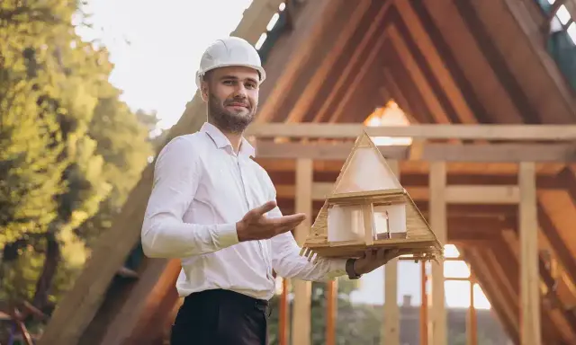 Architekt mit Helm hält Modellhaus, um Holzkonstruktion zu planen. Im Hintergrund Rohbau eines Hauses.
