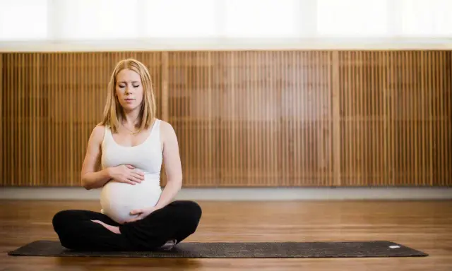 Gravid kvinna med slutna ögon sitter i lotusställning på en yogamatta. Hon håller sin mage, en lugn stund av gravid yoga.
