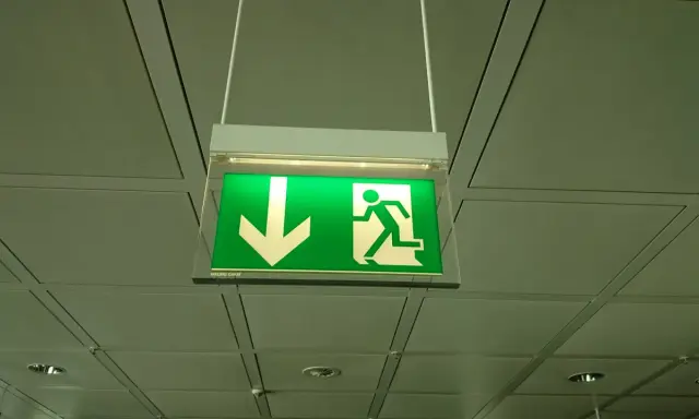 Zielony znak oświetlenia awaryjnego i ewakuacyjnego z ikoną strzałki w dół i biegnącej postaci.