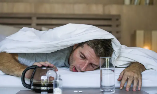 Zmęczony mężczyzna pod kołdrą, obok kawa, woda i tabletki. Może potrzebuje wlewy witaminowe, by odzyskać siły.