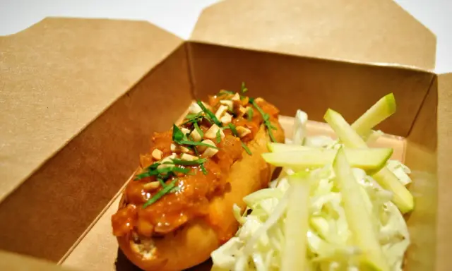 Un delicioso hot dog con salsa de cacahuete y cilantro, acompañado de ensalada de col y manzana, listo para tu delivery.