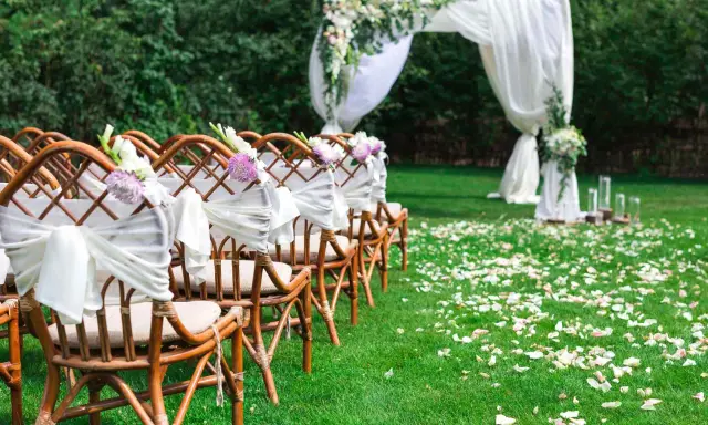 Eleganckie dekoracje na ślub cywilny w plenerze: rzędy krzeseł ozdobionych białymi wstęgami i kwiatami, łuk ślubny z tkaniną i zielenią, ścieżka usypana płatkami róż.