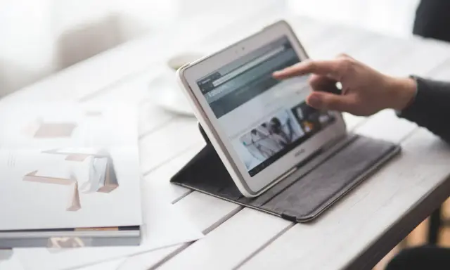 Dłoń wskazuje na ekran tabletu, który wyświetla stronę internetową. Obok leży otwarta książka z grafikami. W tle widać filiżankę. To idealne miejsce do pracy nad projektem, gdzie każdy element, nawet **padding CSS**, ma znaczenie.
