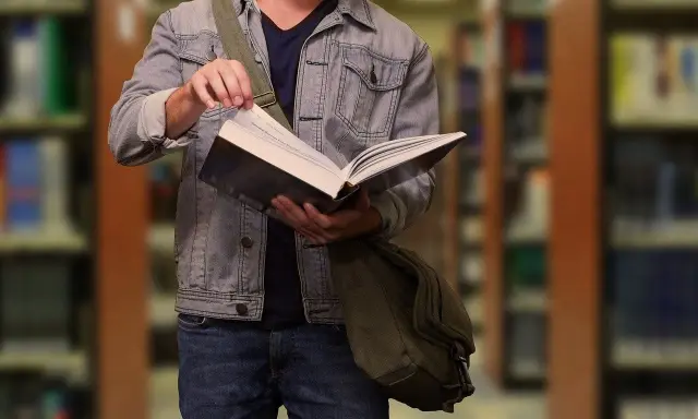 Student w dżinsowej kurtce przegląda książkę w bibliotece. W tle widać regały z książkami.