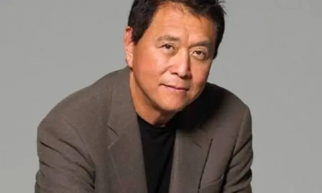 Robert Kiyosaki, autor "Bogatych ojców", w brązowej marynarce i czarnej koszulce, patrzy w obiektyw.