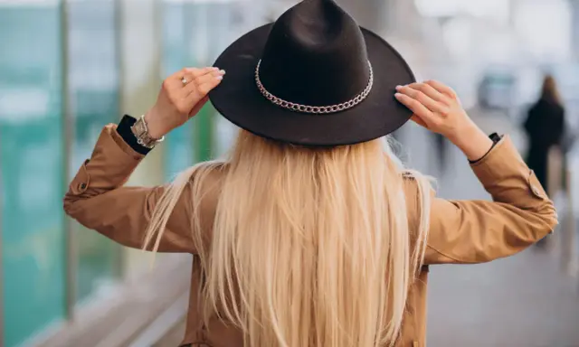 Blondynka w beżowym płaszczu i czarnym kapeluszu z ozdobnym łańcuszkiem. Pokazuje, jak nosić kapelusz, poprawiając go na głowie.