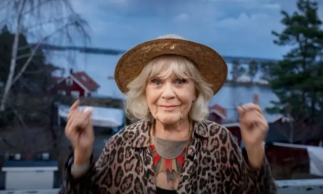 Christina Schollin, med sin karaktäristiska hatt och leopardmönstrade jacka, ser ut att berätta en historia från sina filmer och tv-program.