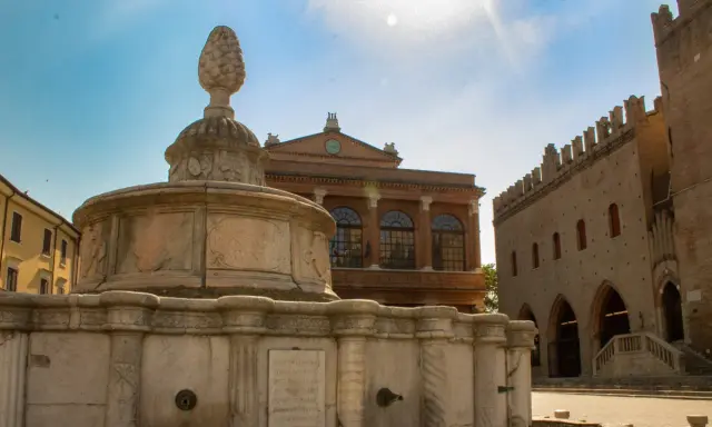 Fontana storica nel centro di Rimini, con edifici antichi sullo sfondo e cielo azzurro.
