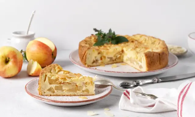 Un délicieux gâteau aux pommes, garni d'amandes effilées, est présenté avec une part déjà coupée. Des pommes fraîches sont disposées à côté.