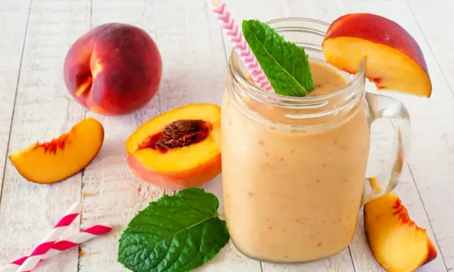 Erfrischender Pfirsich Smoothie im Glas, garniert mit Minze und Pfirsichspalten. Dazu frische Pfirsiche und Trinkhalme.