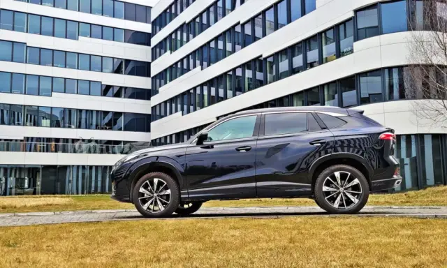 Czarny SUV MG HS, z nowoczesnym designem, zaparkowany przed biurowcem. Jego linia nadwozia sugeruje dynamiczne osiągi i niskie spalanie.