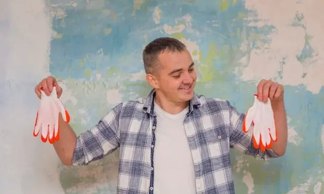 Homme souriant tenant des gants blancs à bouts orange devant un mur texturé. Il semble prêt à peindre, peut-être pour estimer le prix peinture m2 au black.