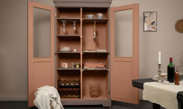 Armoire vitrée avec un relooking meuble 2 couleurs : gris anthracite et rose poudré. Elle contient vaisselle, verres, bouteilles de vin et objets déco.