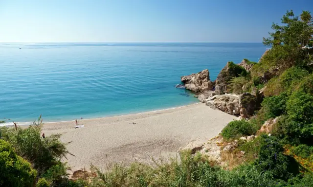 Playa de Burriana: Ein sonniger Strand mit türkisfarbenem Wasser, Felsen und üppiger Vegetation. Einige Menschen genießen die Ruhe.