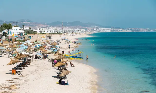 Spiaggia affollata in Marocco con bambini che giocano in acqua e famiglie che si rilassano sotto gli ombrelloni.