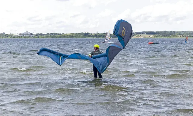 Osoba w żółtym kasku przygotowuje się do **kite surfingu**, trzymając duży, niebiesko-szary latawiec na wzburzonej wodzie.