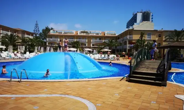 Sole e divertimento in piscina a Maiorca con bambini. Una grande struttura gonfiabile blu al centro, bagnanti e hotel sullo sfondo.