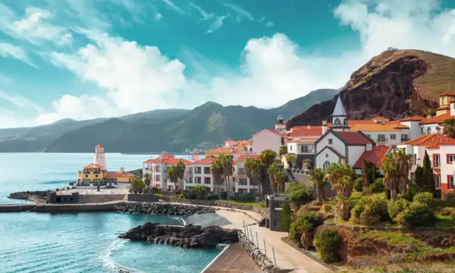 Bunte Häuser mit roten Dächern am Meer, Palmen und Berge im Hintergrund. Ein Leuchtturm wacht über die Küste. Perfektes Klima Portugal.