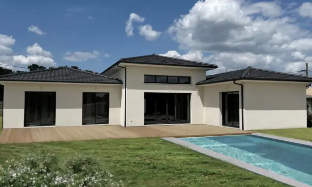 Une maison du sud moderne avec piscine, terrasse en bois et grand jardin sous un ciel bleu nuageux.