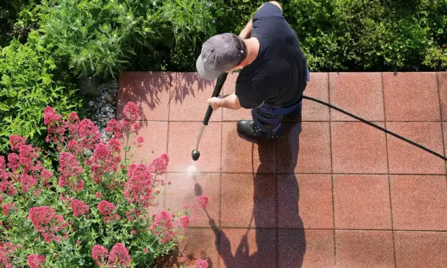 Mężczyzna w czapce z daszkiem czyści kostkę brukową myjką ciśnieniową, obok kwitnące różowe kwiaty i zielone krzewy.