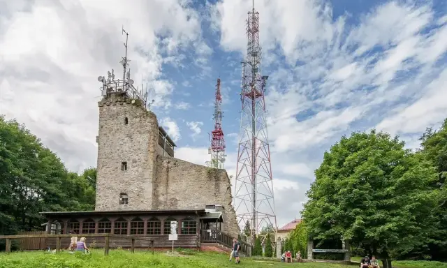 Kamienna wieża i wysokie maszty telekomunikacyjne na szczycie Chełmiec. Ludzie odpoczywają na trawie.