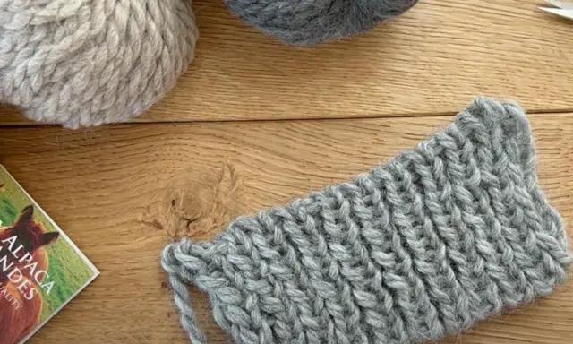 Ovillos de lana gris y beige sobre madera. Un trozo de tejido con puntos de lana muestra la textura.