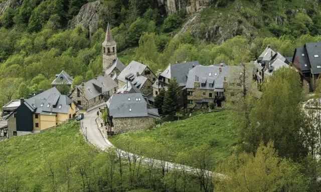 Pueblos del Valle de Arán, con casas de piedra y tejados grises, se asoman entre la vegetación verde y un camino serpenteante.