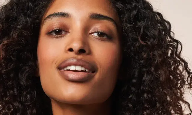 Ein Porträt einer Frau mit voluminösen, dunklen Locken, die die Curly Girl Methode perfektioniert. Ihre Haut strahlt, ihre Augen funkeln.