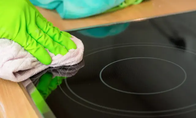 Grüne Handschuhe wischen mit einem Tuch über das Induktionskochfeld, um es zu reinigen.