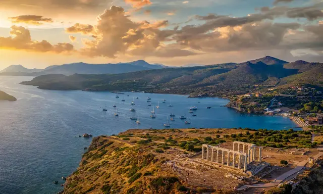 Rovine del Tempio di Poseidone a Capo Sounion, con barche a vela che navigano nel mare blu sotto un cielo dorato al tramonto.