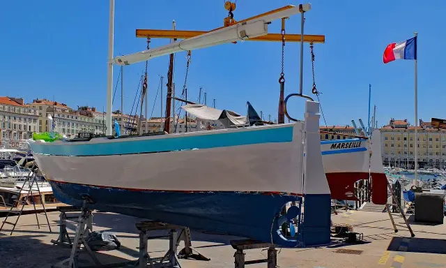 Un bateau hors de l'eau, prêt pour la maintenance, avec le nom "MARSEILLE" peint sur sa coque. Le drapeau français flotte au loin.