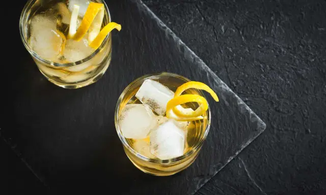 Zwei erfrischende Whiskey Cocktails mit Eis und Zitronenzesten auf dunklem Stein.